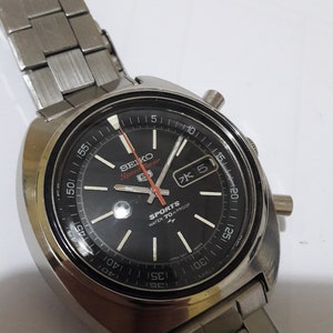 May include: A silver Seiko Speed Timer chronograph wristwatch with a black face, red accents, and a silver metal band. The watch face has white markings and the text "SEIKO Speed Timer", "SPORTS", "WATER 70 m PROOF", and "JAPAN 7031 4005".