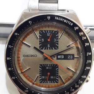 May include: A silver-toned Seiko chronograph wristwatch with a metallic champagne dial, black tachymeter bezel, and stainless steel link bracelet. The watch face features two black sub-dials, orange hands, and a day-date window displaying WED 23.