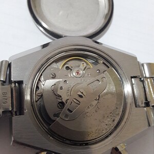 May include: Close-up view of the back of a silver wristwatch with the case open, revealing the inner workings of the watch movement. The watch has a black leather band and a silver face.
