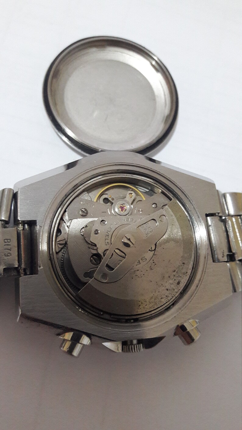 May include: Close-up view of the back of a silver wristwatch with the case open, revealing the inner workings of the watch movement. The watch has a black leather band and a silver face.