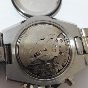 May include: Close-up view of the back of a silver wristwatch with a visible mechanical movement. The watch has a round case and a metal band.