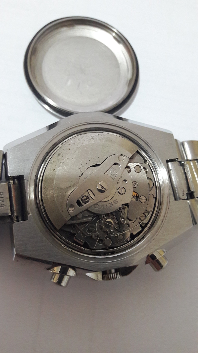 May include: Close-up view of the back of a silver wristwatch with a visible mechanical movement. The watch has a round case and a metal band.