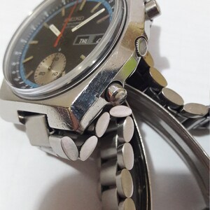 May include: A silver wristwatch with a black face and a blue and white tachymeter scale. The watch has a silver metal band with a push-button clasp.