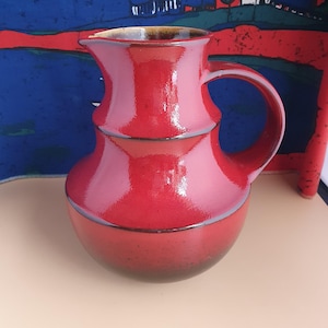 Może przedstawiać: Błyszczący, czerwony ceramiczny dzbanek z uchwytem. Dzbanek ma pękatą podstawę, cylindryczną środkową część i rozszerzoną górę z dzióbkiem. Obręcz i podstawa mają ciemniejszy, brązowawy odcień. Tło przedstawia kolorowy abstrakcyjny obraz.