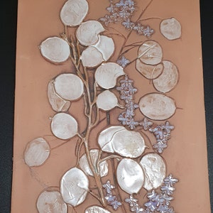Pode incluir: Um azulejo de terracota com um desenho em relevo de um ramo com folhas brancas redondas e flores roxas.