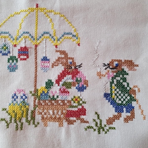 Könnte beinhalten: Kreuzstich-Stickerei mit Osterhasen unter einem gelben Regenschirm. Die Szene zeigt Hasen, Ostereier und hängende Laternen, alles im farbenfrohen, volkstümlichen Stil. Die Stickerei befindet sich auf einem weißen Stoffhintergrund.