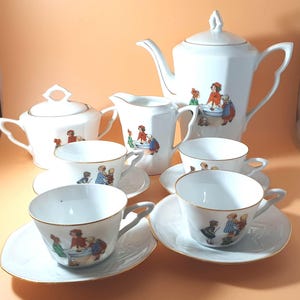 Può includere: Un set da tè in porcellana bianca con bordo dorato e un'illustrazione di bambini. Il set comprende una teiera, un creamer, una zuccheriera e quattro tazze da tè e piattini.