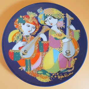 May include: Ceramic plate with a dark blue border, depicting two figures playing stringed instruments. The figures are dressed in colourful, patterned clothing with gold accents. The plate is signed "Bijan Wischild."