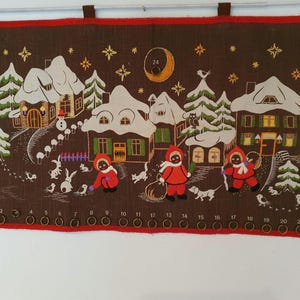 May include: Brown fabric advent calendar depicting a winter village scene. The design includes snow-covered houses, trees, and figures in red coats. The calendar has 24 numbered eyelets along the bottom edge.