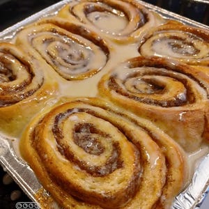 May include: A close-up shot of freshly baked cinnamon rolls in a foil pan. The rolls are golden brown with a swirl of cinnamon and a generous drizzle of white icing. The image is well-lit, showcasing the texture and details of the baked goods.