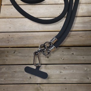 Könnte beinhalten: Schwarzes Seil-Leine mit einem silbernen Metallclip und einem schwarzen Griff. Die Leine liegt auf einer Holzoberfläche.