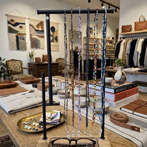 Puede incluir: Una exhibición de accesorios para gafas, que incluye cadenas de gafas con cuentas, exhibidas en un soporte de madera y metal. El soporte contiene un par de gafas de carey. El fondo muestra el interior de una tienda.