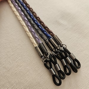 May include: Braided eyeglass chains in cream, lavender, gray, blue, and brown. Each chain has a metal clasp and black rubber loops for securing to glasses. The chains are displayed on a textured, off-white surface.