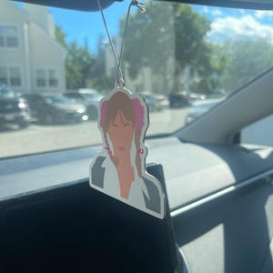 May include: A hanging car air freshener featuring a stylized illustration of a person with light brown hair styled in two braids with pink hair ties, wearing a white top with a dark gray collar, suspended by a thin white string from a rearview mirror.