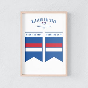 May include: A framed print with the Western Bulldogs Football Club logo and the text "Premiers 1954" and "Premiers 2016." The design features blue, red, and white flags. The frame is a light wood colour.