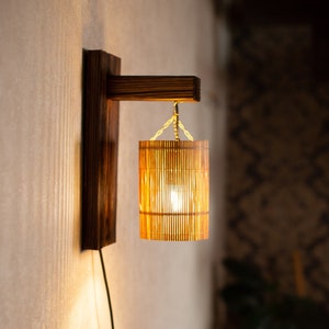 Könnte beinhalten: Eine Wandleuchte mit einem Holzsockel und einem zylindrischen Bambus-Schirm. Das Licht ist eingeschaltet und strahlt ein warmes Licht aus.