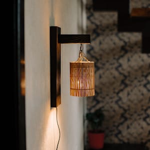 Puede incluir: Una lámpara de pared de madera con una pantalla de bambú colgada de una cadena. La luz está encendida y proyecta un brillo cálido.