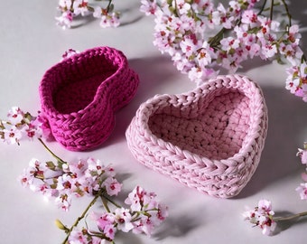 May include: Two crocheted heart-shaped baskets, one pink and one fuchsia, on a white surface with pink flowers.