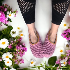 Op de afbeelding: Een paar roze gehaakte pantoffels met gele bies. De pantoffels worden gedragen door iemand die zwarte leggings draagt.