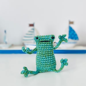 Op de afbeelding: Een groen en blauw gehaakt kikker speelgoed met een brede glimlach en grote ogen. De kikker zit op een wit oppervlak met een wazige achtergrond van speelgoed zeilboten.