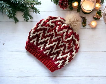 May include: A red and white crocheted beanie with a fluffy pom pom on top. The beanie is lying on a white wooden surface.