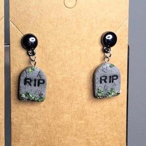 May include: A pair of novelty earrings featuring gray tombstone charms with the letters "RIP" and green moss accents. The earrings have black stud tops and silver-tone chain links.