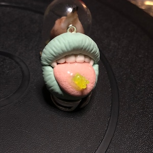 May include: A unique pendant featuring a sculpted mouth with a light green lip color, white teeth, and a pink tongue with a yellow substance. A clear glass dome is above the mouth. The pendant is attached to a silver-colored ring.