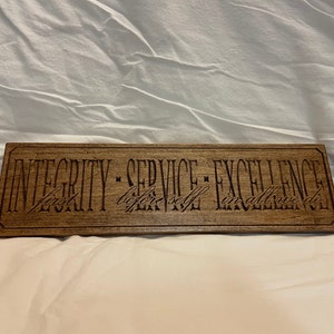 May include: A rectangular wooden sign with the words "INTEGRITY - SERVICE - EXCELLENCE" engraved in a dark font. Below the main text are the words "first, before self, in all we do". The sign is a medium brown color.