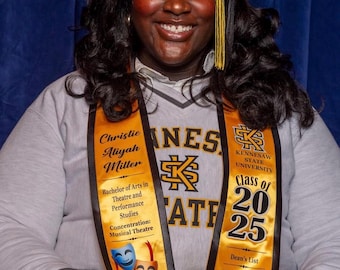 May include: A graduate wearing a black mortarboard cap with a gold tassel and a light gray sweatshirt featuring the Kennesaw State University logo. Two gold graduation stoles are draped over the shoulders, displaying text, theater masks, and an owl.