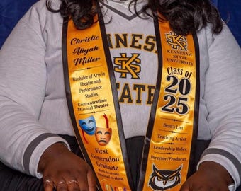 May include: A light gray V-neck sweatshirt with Kennesaw State University branding worn with two gold graduation stoles. The stoles feature black text, the university logo, theater masks, an owl graphic, and the text Class of 2025.