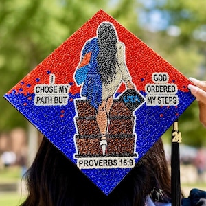 May include: A diamond-shaped graduation cap topper decorated with red and blue rhinestones. It features a figure climbing stairs, with the text: I CHOSE MY PATH BUT GOD ORDERED MY STEPS, UTA, and PROVERBS 16:9. A black tassel with a 24 charm is attached.