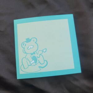 May include: A square notepad with a light blue interior and a teal border. A teal illustration of a bear wearing a hat and bow tie is in the lower left corner. The notepad is on a dark surface.