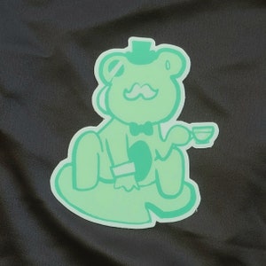 May include: A light green sticker featuring a cartoon frog wearing a top hat, bow tie, and mustache, holding a teacup. The frog is sitting on a lily pad, with a light green outline against a dark background.