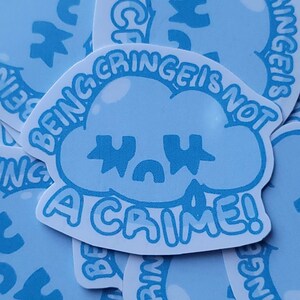 May include: Light blue sticker with a cloud-shaped design and the text "BEING CRINGE IS NOT A CRIME!". The sticker has a white border and a cartoonish aesthetic. Several stickers are visible.