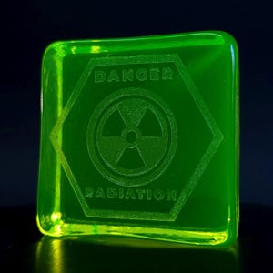 Op de afbeelding: Een lichtgevend groen vierkant met de woorden "DANGER RADIATION" in het oppervlak gegraveerd. Het ontwerp bevat een gevaarsymbool in een zeshoek. Het object heeft een licht afgeronde vorm en een donkere achtergrond.