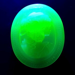 May include: A glowing green glass oval with a detailed relief of a classical figure. The glass has a textured surface and is set against a dark blue background. The oval shape is approximately 4 inches in diameter.