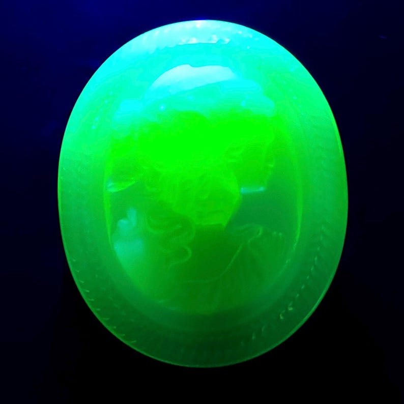 May include: A glowing green glass oval with a detailed relief of a classical figure. The glass has a textured surface and is set against a dark blue background. The oval shape is approximately 4 inches in diameter.