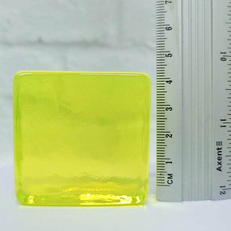 Puede incluir: Un objeto cuadrado de vidrio amarillo ne&oacute;n con una superficie texturizada. Una regla con marcas de cent&iacute;metros se coloca junto al objeto de vidrio. El objeto de vidrio mide aproximadamente 5 cm de alto.