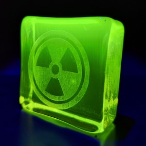 Op de afbeelding: Een gloeiend, neon groen, vierkant glazen blok met een stralingssymbool op het oppervlak gegraveerd. Het symbool is een cirkel met drie uitstralende bladen. Het blok is van binnenuit verlicht, wat een levendig, opvallend effect creëert. Afmetingen ca. 5 cm x 5 cm.