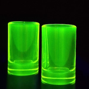 Könnte beinhalten: Zwei zylindrische Schnapsgläser aus durchscheinendem Glas, die ein leuchtendes, neon-grünes Licht abgeben. Die Gläser haben eine glatte, klare Oberfläche und einen dicken, massiven Boden und stehen auf einem dunklen, schwarzen Hintergrund.