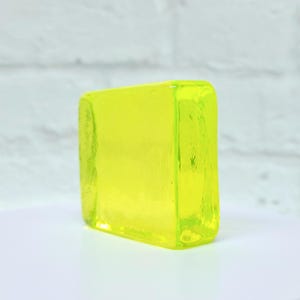 Puede incluir: Un bloque rectangular transl&uacute;cido de vidrio amarillo brillante. Los bordes son ligeramente redondeados y el vidrio tiene un aspecto liso y pulido. El bloque est&aacute; colocado sobre una superficie blanca contra una pared de ladrillos blancos.