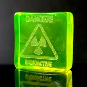 Op de afbeelding: Een lichtgevend groen, vierkant object met het woord "DANGER!" boven een radioactief symbool. Het woord "RADIOACTIVE" staat onder het symbool. Het object wordt weerspiegeld op een zwart oppervlak.