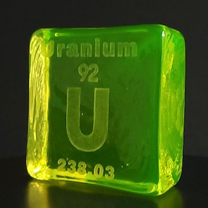 May include: A translucent, square-shaped block of green glass with the word "Uranium" etched at the top. The number "92" and the letter "U" are etched below, with "238.03" at the bottom. The edges of the block are illuminated with a yellow glow.