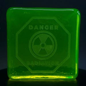 Op de afbeelding: Een vierkant, doorschijnend, neon groen glazen blok met geëtste tekst en een symbool. De tekst luidt "DANGER RADIATION" rond een stralingsgevaarsymbool. Het blok wordt van onderaf verlicht, waardoor een gloei-effect ontstaat.