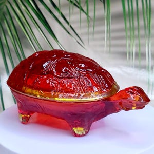 May include: A translucent red glass turtle figurine with a detailed, textured shell pattern. The object features a rounded, domed back, four short legs, and a small head extending forward, all crafted from a single piece of vibrant red glass.