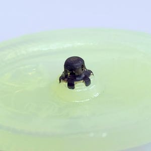 May include: Close-up of a pale green glass object with a dark metal cap. The cap has a textured surface and is centered on the glass. The glass has a slightly uneven surface. The background is a soft, blurred white.