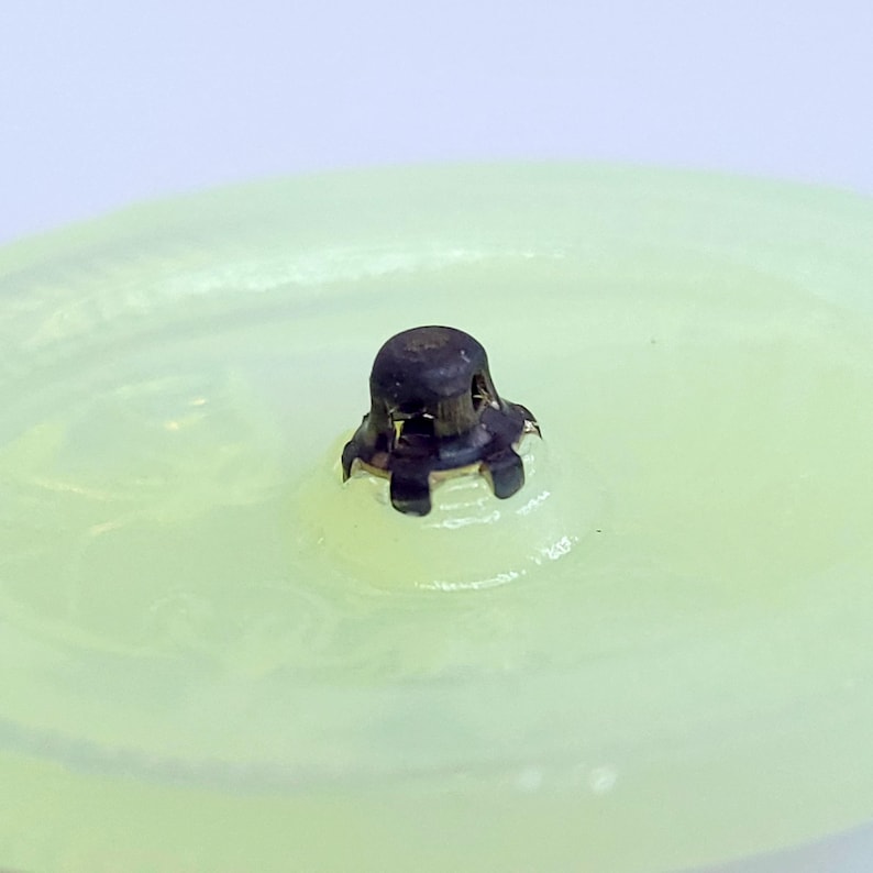 May include: Close-up of a pale green glass object with a dark metal cap. The cap has a textured surface and is centered on the glass. The glass has a slightly uneven surface. The background is a soft, blurred white.