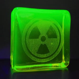 Op de afbeelding: Een gloeiend, vierkant, neon groen object met afgeronde randen. Het object heeft een stralingssymbool in het oppervlak geëtst. De donkere achtergrond benadrukt de heldergroene kleur van het object.