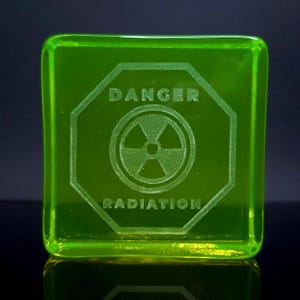 Op de afbeelding: Een vierkant, transparant, neon groen glazen blok met de woorden "DANGER RADIATION" in het oppervlak geëtst. Het ontwerp bevat een stralingssymbool binnen een achthoekige rand.