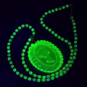 May include: A necklace featuring a series of small, round, translucent beads and a large, oval-shaped pendant. The pendant displays a relief profile of a person's head. The entire piece glows with a bright, neon green light against a dark background.